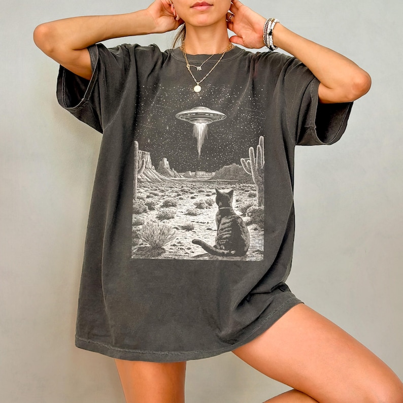 May include: A dark gray oversized t-shirt with a graphic print of a cat looking up at a UFO in a desert landscape.