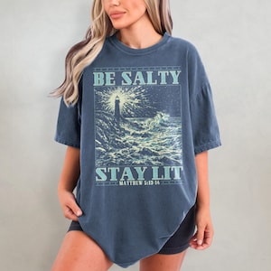 May include: A dark blue oversized t-shirt with a graphic print of a lighthouse in a stormy sea. The text "BE SALTY STAY LIT" is printed on the shirt, with the scripture reference "MATTHEW 5:13-14" below.