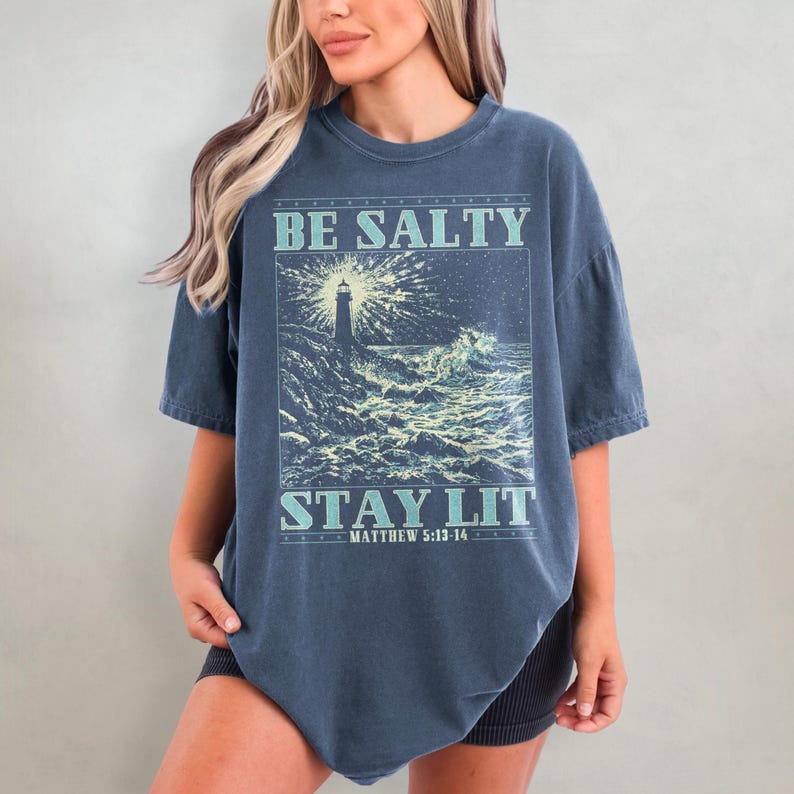 May include: A dark blue oversized t-shirt with a graphic print of a lighthouse in a stormy sea. The text "BE SALTY STAY LIT" is printed on the shirt, with the scripture reference "MATTHEW 5:13-14" below.