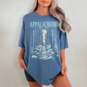 May include: A blue oversized t-shirt with a graphic print of a forest path and the text "Appalachians Est. 1937".