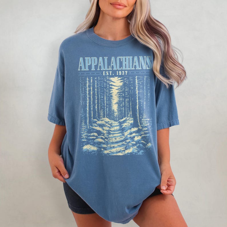 May include: A blue oversized t-shirt with a graphic print of a forest path and the text "Appalachians Est. 1937".