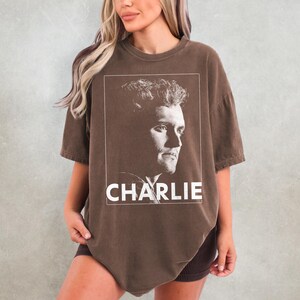 May include: Brown t-shirt with a portrait of a man in a white rectangular frame. The name "CHARLIE" is printed in white block letters below the portrait. The shirt has short sleeves and a relaxed fit.