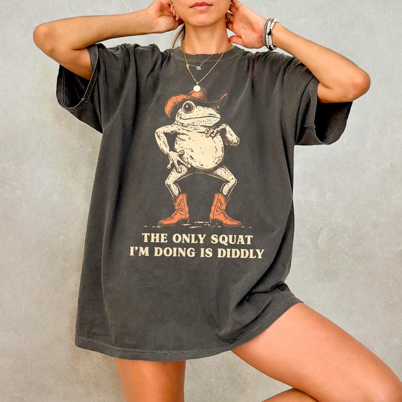 May include: Dark grey t-shirt featuring a cartoon frog wearing a cowboy hat and boots. The frog is squatting with its arms at its sides. The text "THE ONLY SQUAT I'M DOING IS DIDDLY" is printed below the frog.