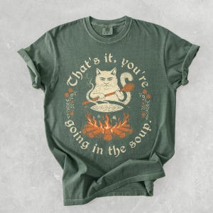 May include: Green t-shirt with a humorous graphic of a cat stirring a pot over a fire, with the text "That's it, you're going in the soup." The design includes orange flames, a wooden spoon, and floral accents.