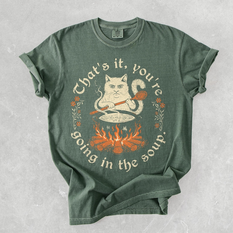 May include: Green t-shirt with a humorous graphic of a cat stirring a pot over a fire, with the text "That's it, you're going in the soup." The design includes orange flames, a wooden spoon, and floral accents.