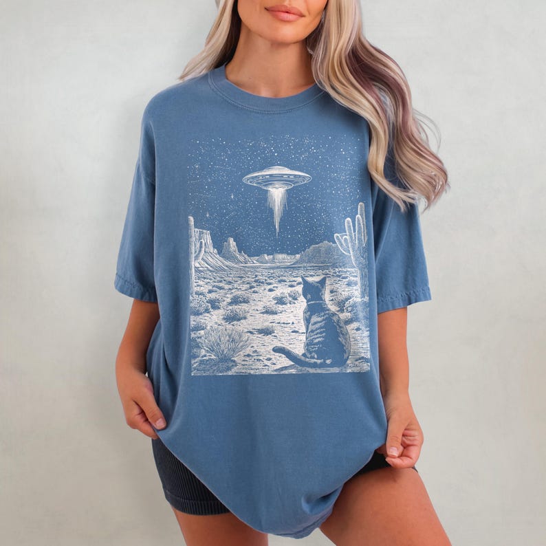 May include: A blue t-shirt with a white graphic of a cat looking up at a UFO in a desert landscape. The graphic also includes stars and cacti.