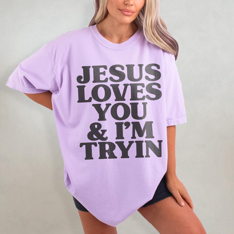 May include: A lavender oversized t-shirt with the text "JESUS LOVES YOU & I'M TRYIN" printed in black.