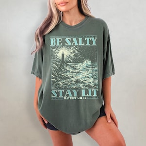 May include: A green oversized t-shirt with a graphic of a lighthouse and waves. The text on the shirt reads "Be Salty Stay Lit Matthew 5:13-14".