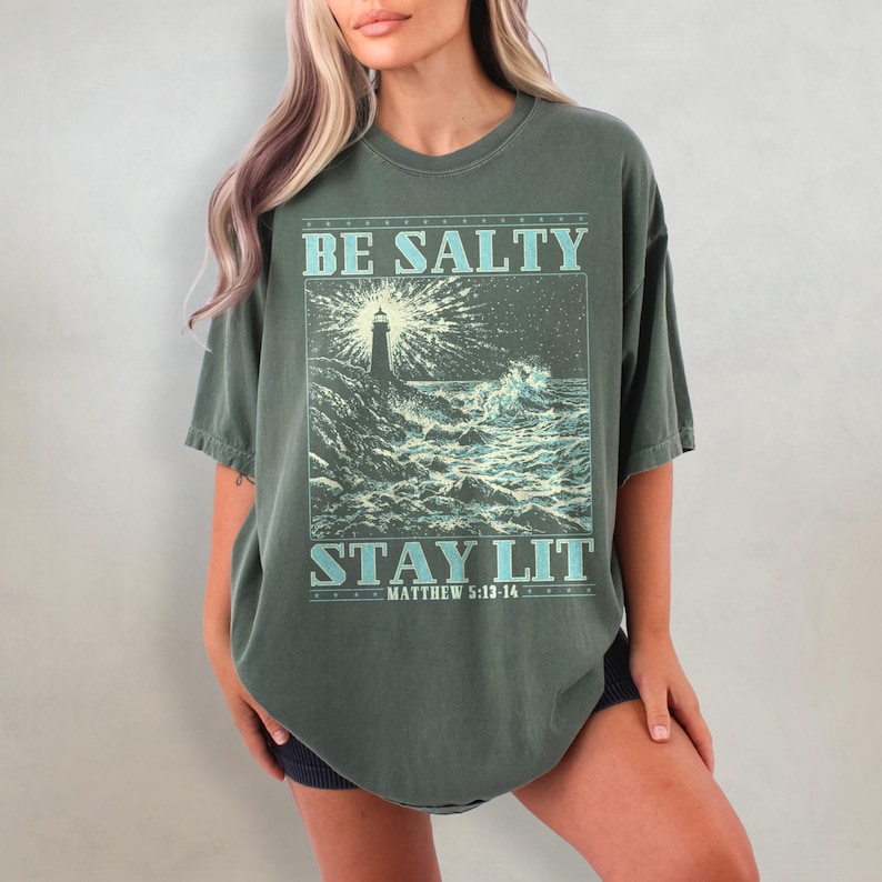 May include: A green oversized t-shirt with a graphic of a lighthouse and waves. The text on the shirt reads "Be Salty Stay Lit Matthew 5:13-14".