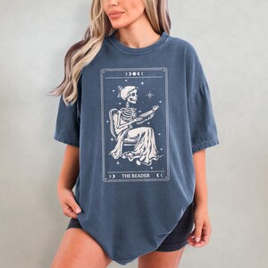 Op de afbeelding: Een marineblauw T-shirt met een witte illustratie van een skelet dat een boek leest, omlijst door een rechthoek met hemelse accenten. De tekst "THE READER" staat onderaan het frame.