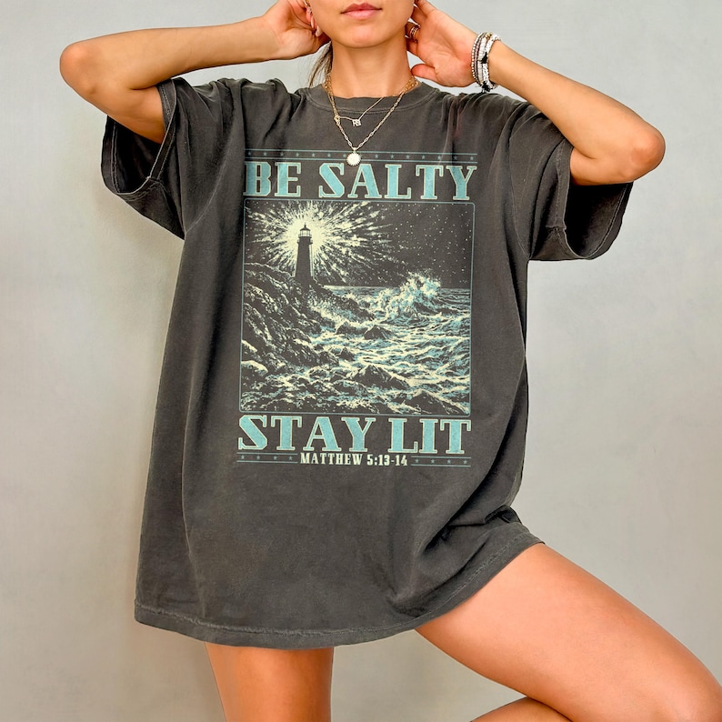 May include: A dark gray oversized t-shirt with a graphic print of a lighthouse in a stormy sea. The text "BE SALTY STAY LIT" is printed in blue on the shirt. The text "MATTHEW 5:13-14" is printed in smaller font below the graphic.