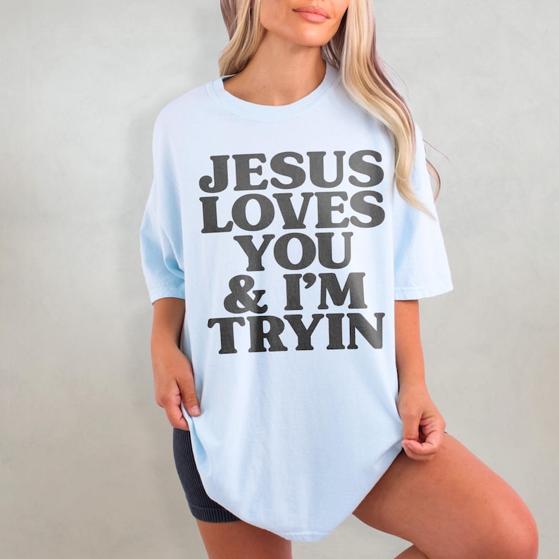 May include: A light blue oversized t-shirt with the text "JESUS LOVES YOU & I'M TRYIN" printed in black.