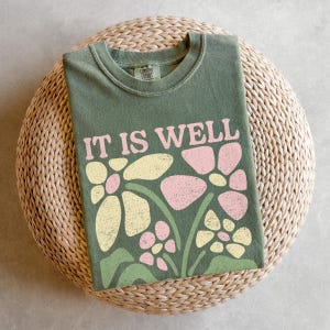 May include: Olive green t-shirt with the phrase "IT IS WELL" in pink letters. The shirt features a floral design in yellow and pink. The t-shirt is folded and resting on a woven, circular surface.