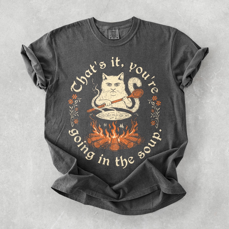 May include: Dark gray t-shirt featuring a whimsical illustration of a cat stirring a pot over a fire. The text reads "That's it, you're going in the soup." The design is encircled by floral accents.