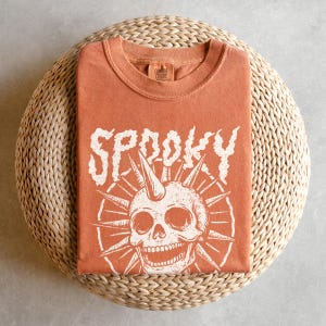 May include: An orange t-shirt with the word "SP00KY" in white, stylized lettering. Below the word is a white skull graphic with a horn and spiky sunburst design. The shirt is folded on a woven, circular surface.