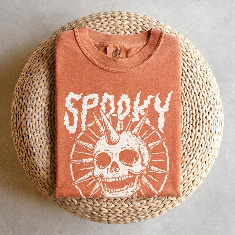 May include: An orange t-shirt with the word "SP00KY" in white, stylized lettering. Below the word is a white skull graphic with a horn and spiky sunburst design. The shirt is folded on a woven, circular surface.