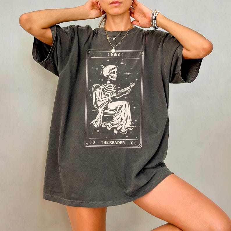 Op de afbeelding: Donkergrijs t-shirt met een witte illustratie van een lezend skelet, zittend in een stoel. Het ontwerp is omlijst met een rand en de tekst "THE READER" staat onderaan. Het shirt heeft korte mouwen en een relaxte pasvorm.