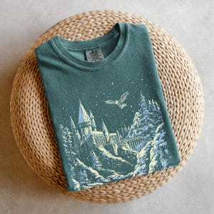 Puede incluir: Una camiseta de color verde azulado con un gráfico de un castillo, montañas nevadas y un búho volando bajo un cielo estrellado. El diseño es en blanco y azul claro. La camiseta está doblada sobre una superficie circular tejida.