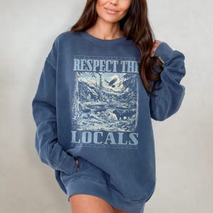 May include: A blue oversized sweatshirt with a graphic print of a mountain scene, an eagle, and the text "Respect the Locals".