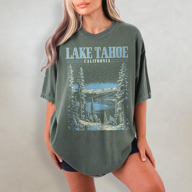 May include: A green oversized t-shirt with a vintage-style graphic of Lake Tahoe, California. The graphic features a snowy mountain scene with a lake and pine trees.