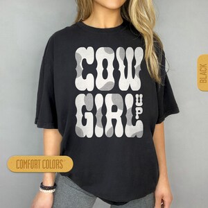 May include: Black t-shirt with a white graphic that says "COWGIRL UP!" in a retro font with a cow print pattern.