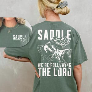 May include: Sage green t-shirt with a white graphic of a cowboy riding a horse, and the text "Saddle Up, We're Following The Lord." The front of the shirt has the text "Saddle Up" in a script font.
