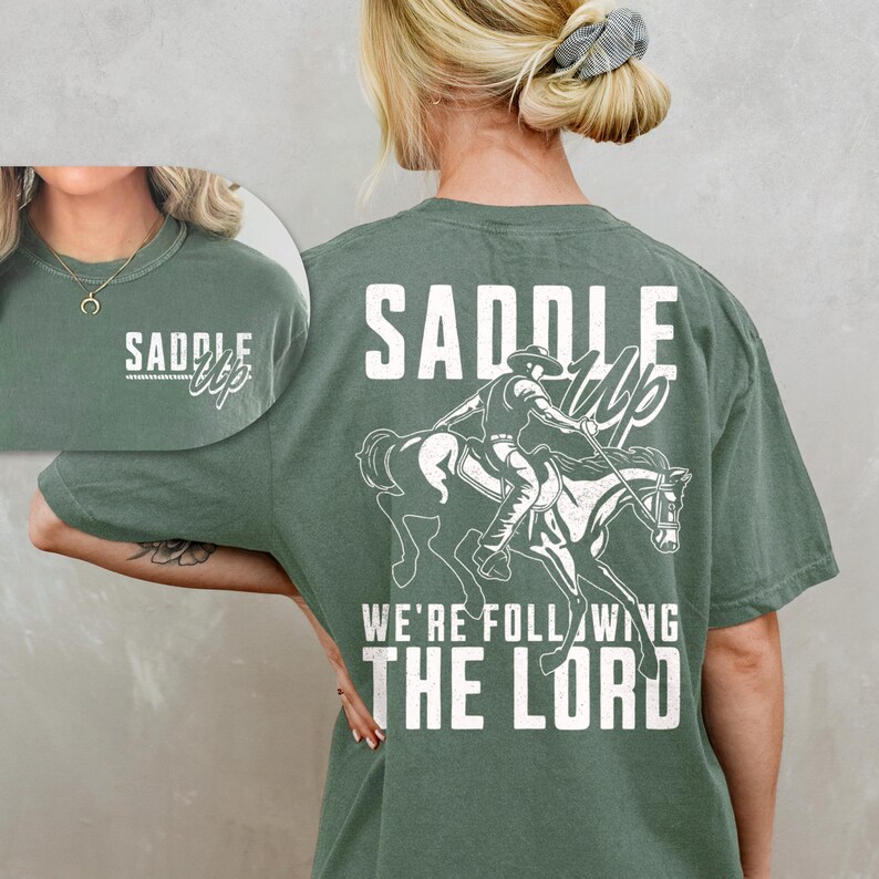 May include: Sage green t-shirt with a white graphic of a cowboy riding a horse, and the text "Saddle Up, We're Following The Lord." The front of the shirt has the text "Saddle Up" in a script font.
