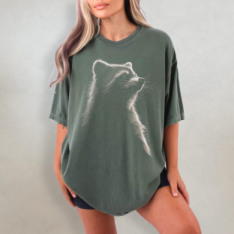 May include: An oversized green t-shirt featuring a white graphic of a raccoon looking upwards. The raccoon is in profile and has a detailed, realistic design.