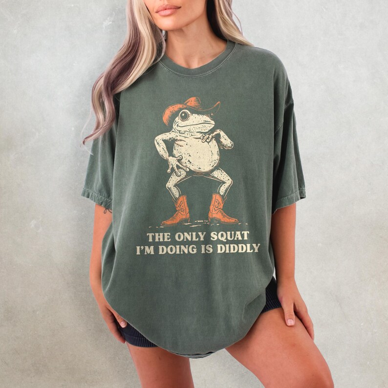 May include: Green t-shirt featuring a cartoon frog wearing a cowboy hat and boots. The frog is in a squatting pose. The text "THE ONLY SQUAT I'M DOING IS DIDDLY" is printed below the frog.