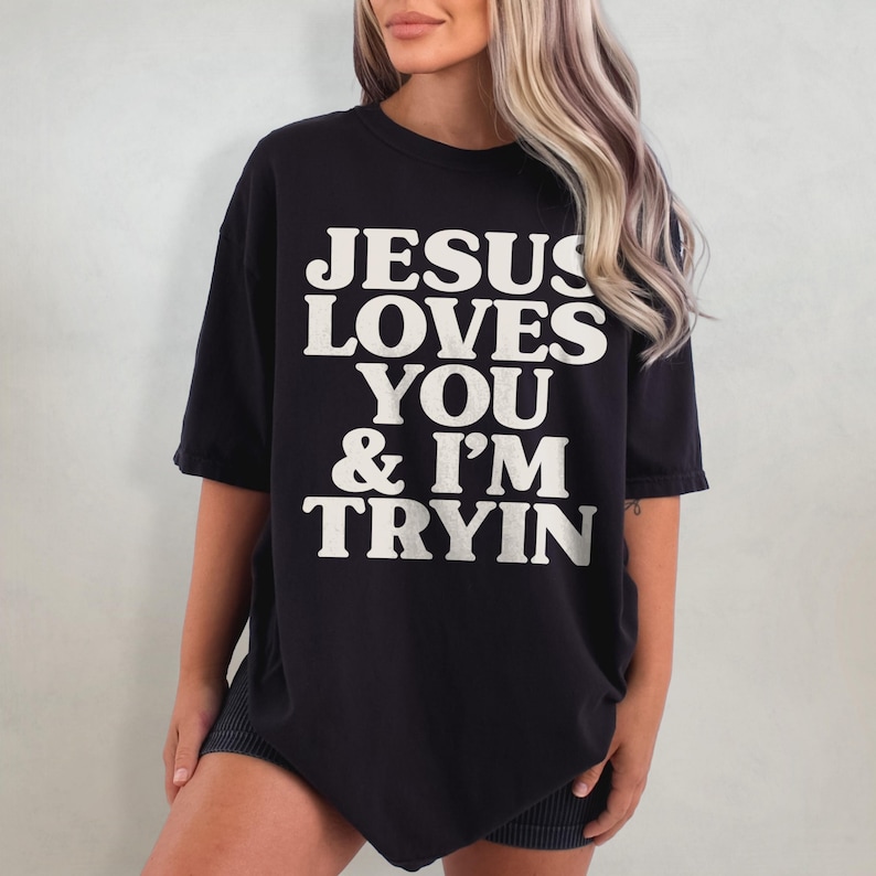 May include: Black oversized t-shirt with white text that reads "JESUS LOVES YOU & I'M TRYIN"