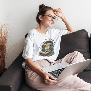 May include: A white t-shirt with a graphic of a cat reading a book and the text "One More Chapter". The cat is wearing a blue and white space suit. The person wearing the shirt is sitting on a couch with a laptop.