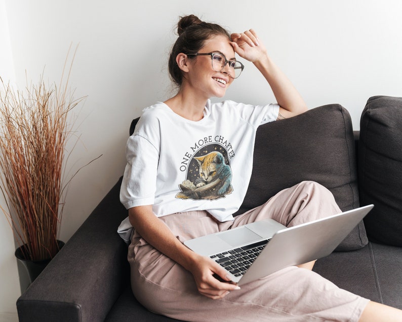 May include: A white t-shirt with a graphic of a cat reading a book and the text "One More Chapter". The cat is wearing a blue and white space suit. The person wearing the shirt is sitting on a couch with a laptop.