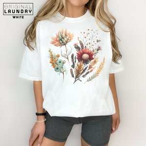 May include: White t-shirt with a floral print featuring a variety of flowers in shades of orange, red, and yellow.