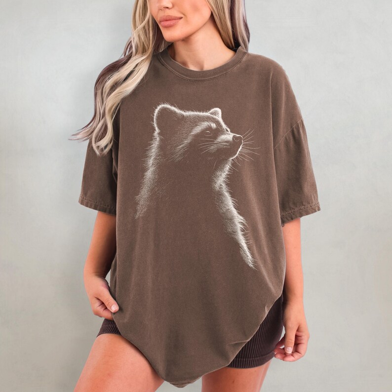 May include: An oversized brown t-shirt featuring a white graphic of a raccoon looking to the right.