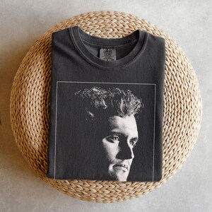 May include: Charcoal grey t-shirt featuring a black and white portrait of a man in a white frame. The shirt is folded on a circular woven mat. The t-shirt has a crew neck.