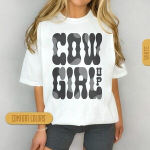 May include: White t-shirt with a black and grey graphic that says "COWGIRL UP" in a retro font.