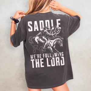 May include: Dark gray t-shirt with a white graphic of a cowboy riding a horse, and the text "SADDLE UP" above and "WE'RE FOLLOWING THE LORD" below. The shirt has short sleeves and a relaxed fit.
