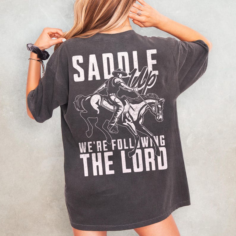 May include: Dark gray t-shirt with a white graphic of a cowboy riding a horse, and the text "SADDLE UP" above and "WE'RE FOLLOWING THE LORD" below. The shirt has short sleeves and a relaxed fit.