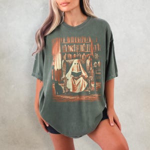 Op de afbeelding: Donkergroen t-shirt met een afbeelding van een geest die in een bibliotheek leest. Het ontwerp heeft een vintage, distressed look met oranje en bruine tinten. Er is ook een zwarte kat aanwezig. Het t-shirt heeft een relaxte pasvorm.