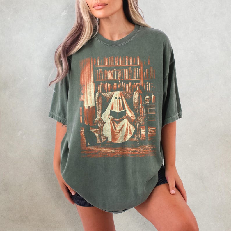 Op de afbeelding: Donkergroen t-shirt met een afbeelding van een geest die in een bibliotheek leest. Het ontwerp heeft een vintage, distressed look met oranje en bruine tinten. Er is ook een zwarte kat aanwezig. Het t-shirt heeft een relaxte pasvorm.