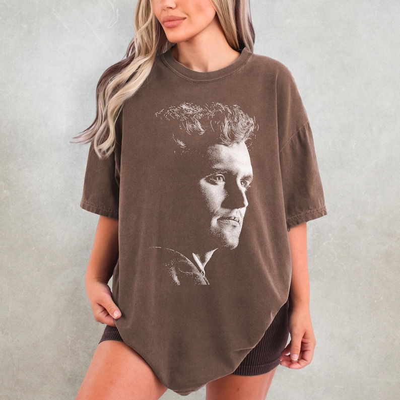 May include: Brown t-shirt featuring a black and white portrait of a man's face. The shirt has short sleeves and a relaxed fit. The image is a close-up of the man's profile, with detailed facial features.