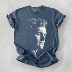 May include: A blue t-shirt featuring a black and white portrait of a man. The portrait is a side profile of the man's face. The shirt has short sleeves and a crew neck.