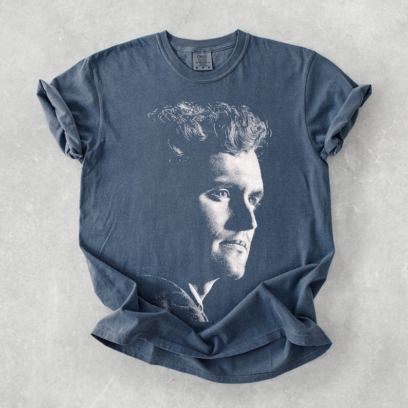 May include: A blue t-shirt featuring a black and white portrait of a man. The portrait is a side profile of the man's face. The shirt has short sleeves and a crew neck.