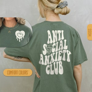 K&ouml;nnte beinhalten: Ein gr&uuml;nes T-Shirt mit dem Text "Anti Social Anxiety Club" in Wei&szlig;, mit einem tropfenden Smiley-Herz-Motiv. Das Shirt ist von Comfort Colors.