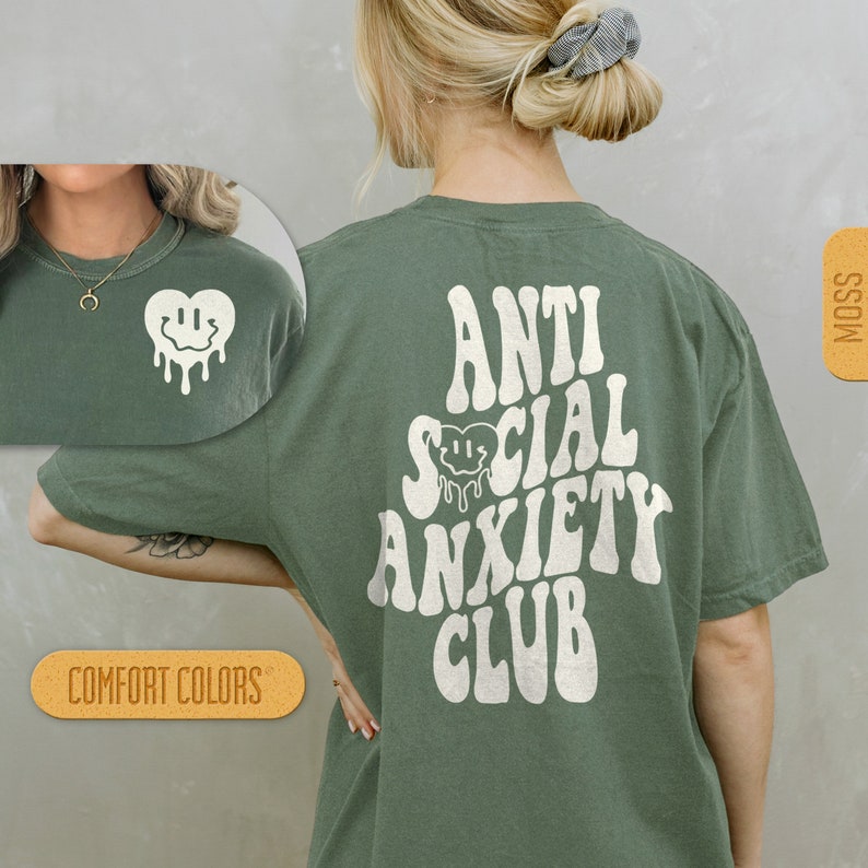 K&ouml;nnte beinhalten: Ein gr&uuml;nes T-Shirt mit dem Text "Anti Social Anxiety Club" in Wei&szlig;, mit einem tropfenden Smiley-Herz-Motiv. Das Shirt ist von Comfort Colors.