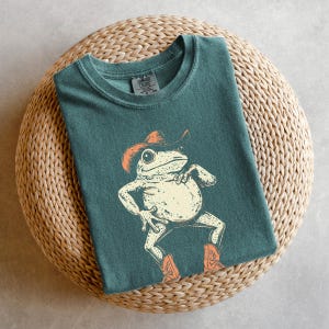 May include: Teal-colored t-shirt featuring a cartoon frog wearing a cowboy hat and boots. The frog is in a dancing pose with its arms and legs spread out. The shirt is folded on a woven, circular surface.
