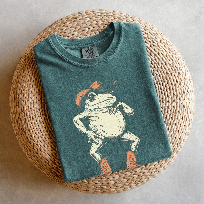 May include: Teal-colored t-shirt featuring a cartoon frog wearing a cowboy hat and boots. The frog is in a dancing pose with its arms and legs spread out. The shirt is folded on a woven, circular surface.