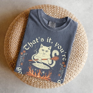 May include: A dark gray t-shirt with the text "That's it, you're done." and a cartoon cat cooking over a fire. The cat has an angry expression and is holding a wooden spoon. The shirt is folded on a woven mat.