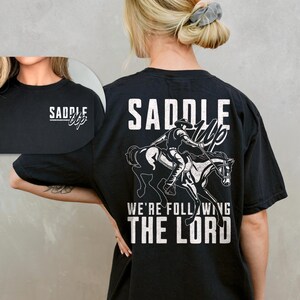 May include: Black t-shirt with a white graphic of a cowboy riding a horse, with the words "SADDLE UP" and "WE'RE FOLLOWING THE LORD". The front of the shirt has the words "SADDLE UP" in white.