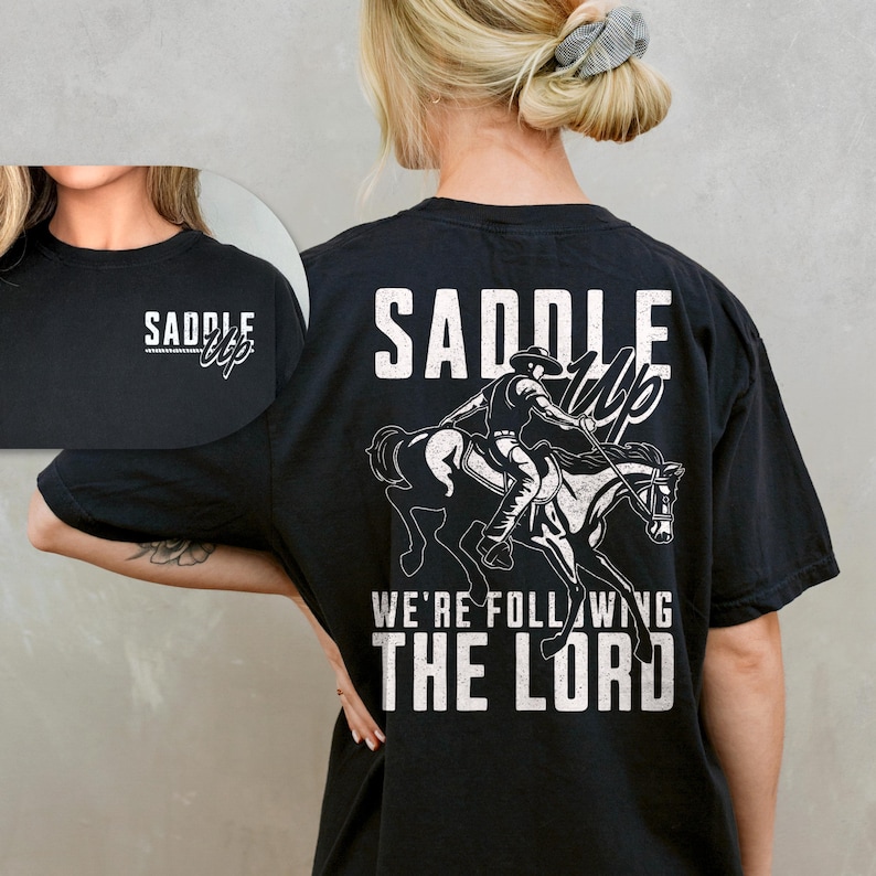May include: Black t-shirt with a white graphic of a cowboy riding a horse, with the words "SADDLE UP" and "WE'RE FOLLOWING THE LORD". The front of the shirt has the words "SADDLE UP" in white.